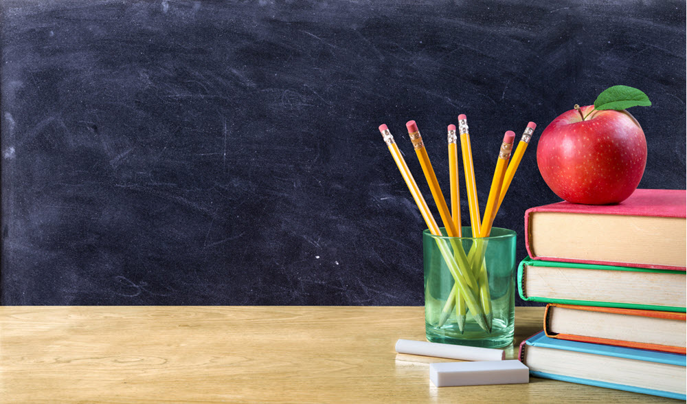 A school desk with chalk, a glass holding 6 pencils and a stack of 4 books on top. An apple is on top of the stack of books. 